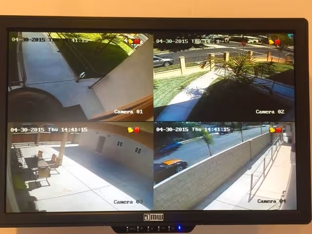 Security camera monitor displaying four different outdoor views around a facility, including a walkway with stairs, a gated entrance with plants, a patio area with chairs and tables, and a sidewalk with a ramp and parked car.