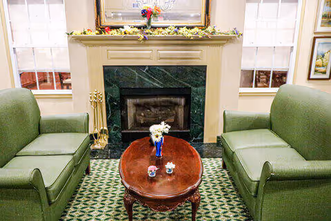 A cozy living room area with two green upholstered armchairs facing each other across a wooden coffee table. The coffee table has a small vase with flowers and two small decorative items. Behind the table is a fireplace with a green marble surround and a mantel decorated with flowers and a framed mirror above it. Two windows with white blinds flank the fireplace.