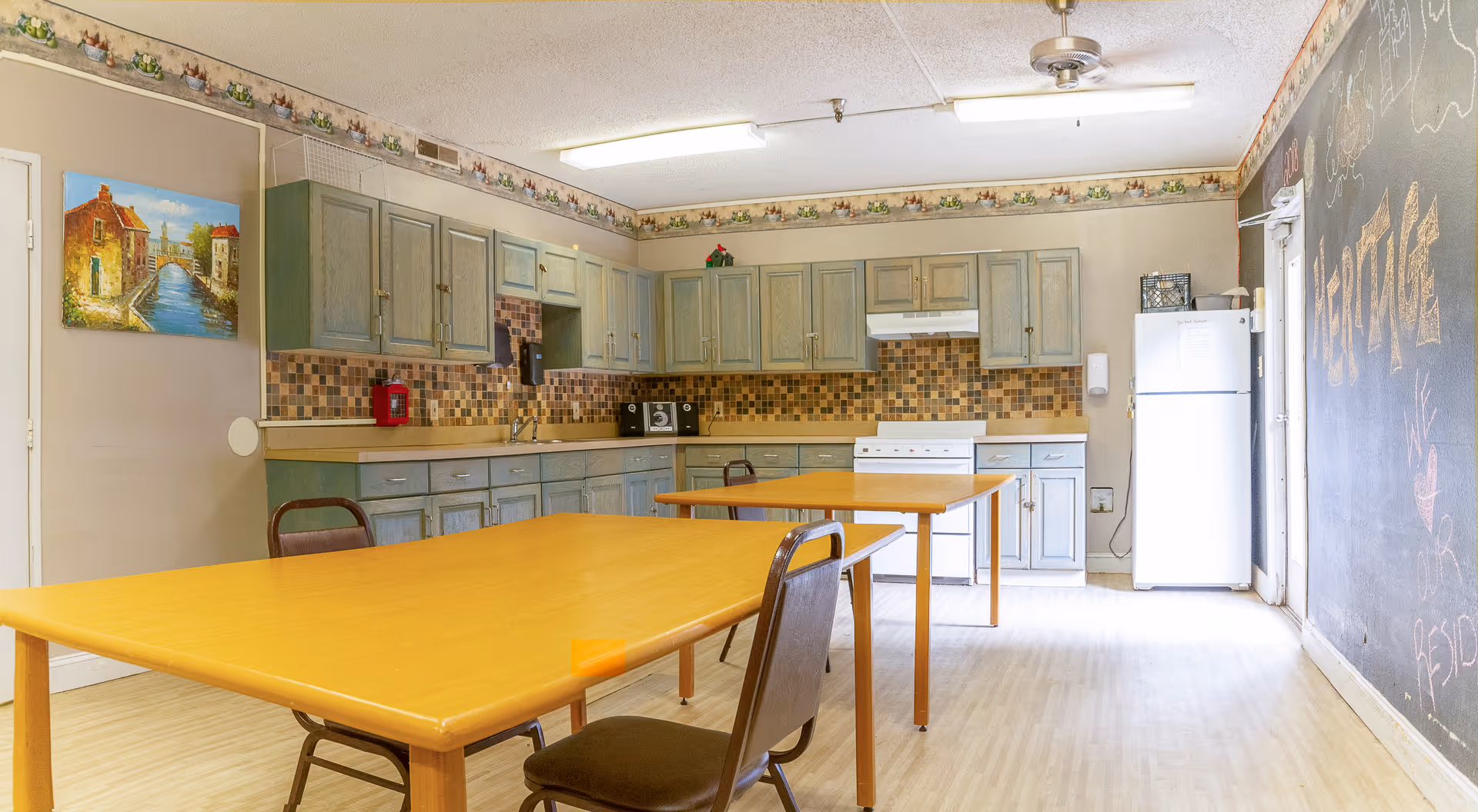 A kitchen area in a senior living facility with light wood tables and chairs, blue-gray cabinets, a white stove, a white refrigerator, and a tiled backsplash. There is a painting on the left wall and a large chalkboard wall on the right with some writing and drawings. The room is well-lit with fluorescent ceiling lights.