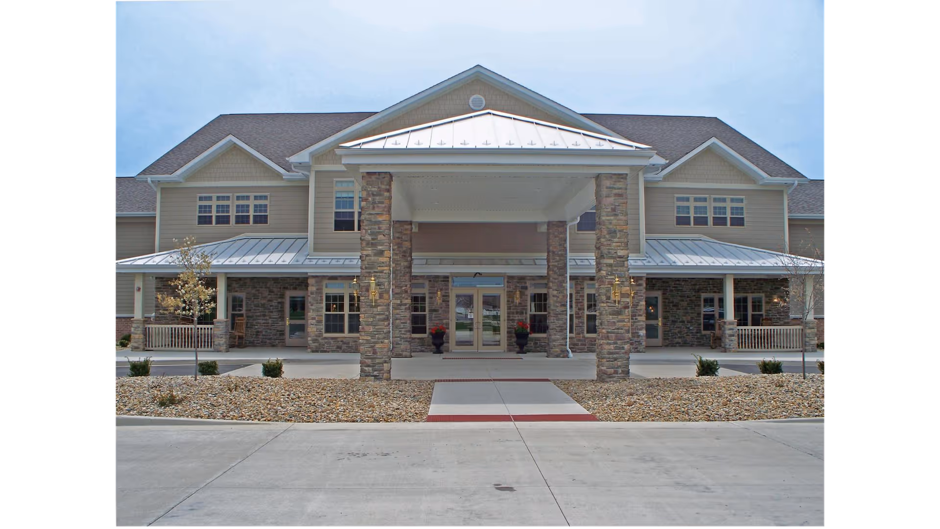 Front exterior view of a two-story building with a covered entrance supported by stone pillars, beige siding, multiple windows, and a paved driveway leading up to the entrance.