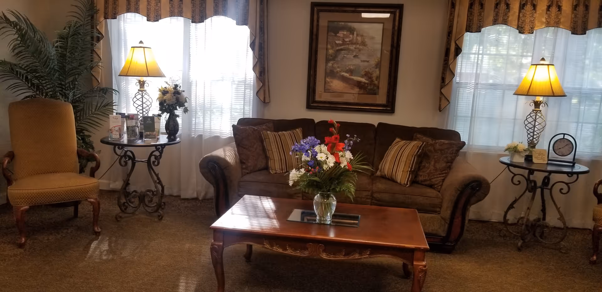A cozy living room area with a brown sofa adorned with striped and patterned cushions, a wooden coffee table with a floral arrangement in a glass vase, two round side tables each with a lamp and decorative items, two upholstered armchairs, and large windows with sheer white curtains and patterned valances.