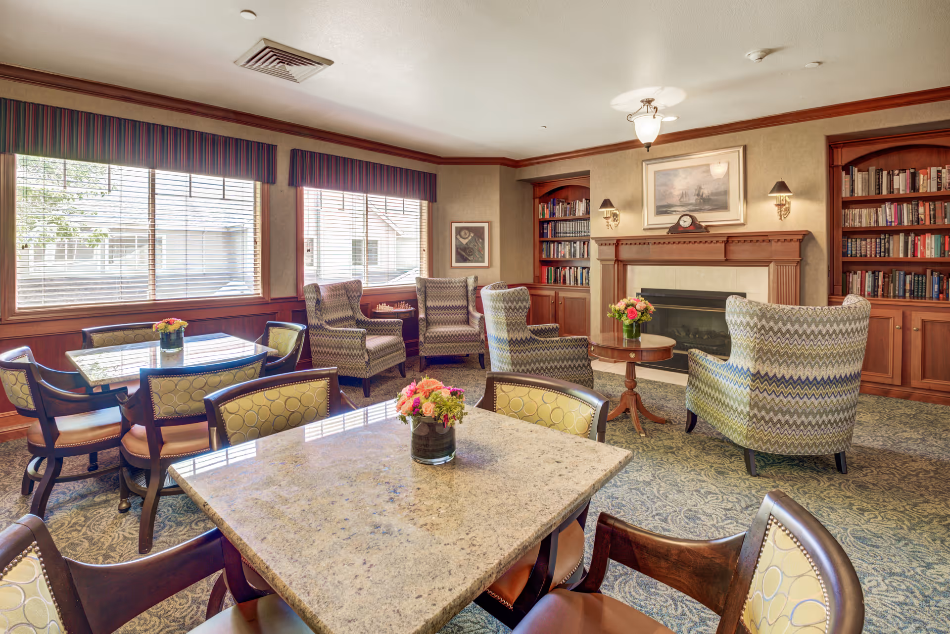 A cozy common area in a senior living facility featuring a fireplace with a mantel clock and framed artwork above it, built-in bookshelves filled with books on either side, several patterned armchairs arranged around a small round table with a flower vase, and two square tables with chairs and flower vases near large windows with blinds and striped valances.