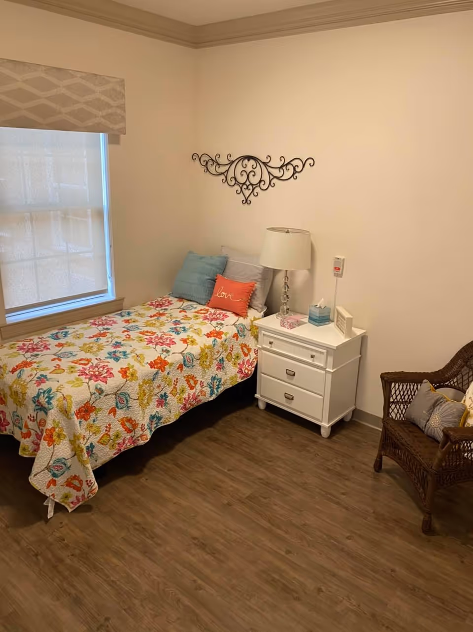 Small furnished bedroom with a single bed in a floral quilt, a white nightstand and lamp, wicker chair, and wall decor.