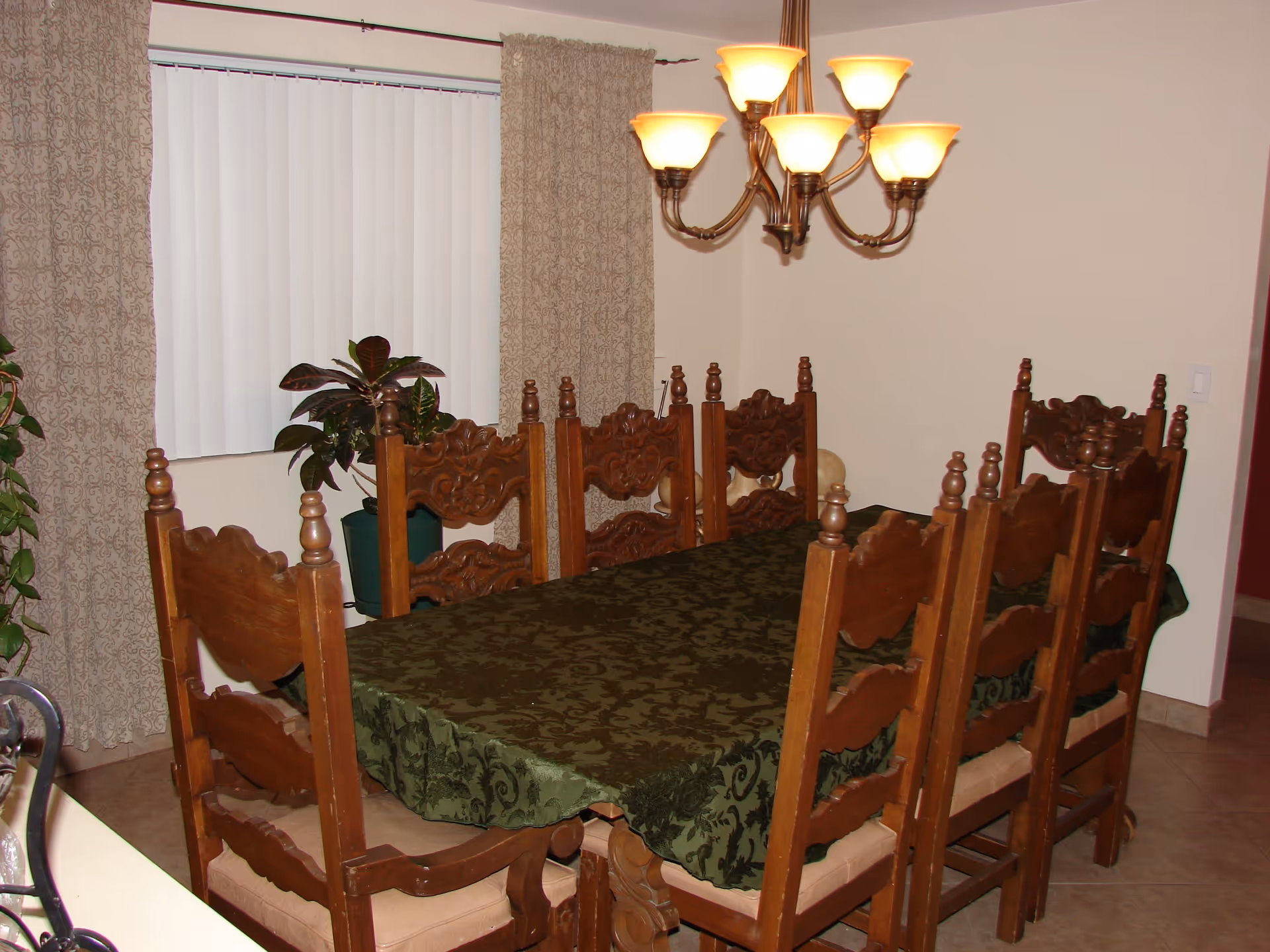 Dining room with a rectangular wooden table covered with a dark green patterned tablecloth, surrounded by eight carved wooden chairs with beige cushions. A chandelier with six lights hangs above the table. There are two potted plants near a window with vertical blinds and patterned curtains.
