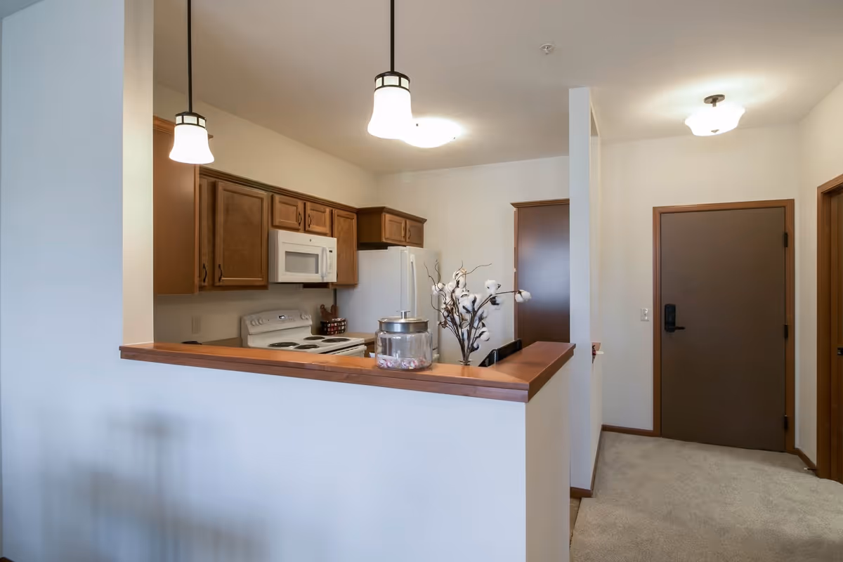 Interior view of a small kitchen and entryway with wood cabinets, a pass-through counter, and pendant lights.