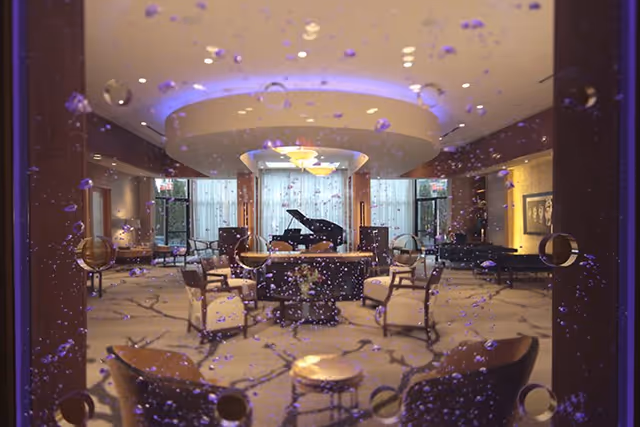 View through a glass wall with water droplets into a spacious, well-lit lounge area featuring multiple armchairs arranged around small tables, a grand piano in the background, and large windows with sheer curtains allowing natural light to fill the room.