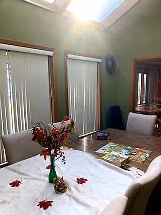 A dining area with a wooden table covered by a white tablecloth decorated with red leaves. A green vase with autumn-themed flowers is placed in the center of the table. There are beige upholstered chairs around the table. Vertical blinds cover the windows, and sunlight streams in from a skylight above. A mirror and some books or puzzles are visible on the table and in the background.