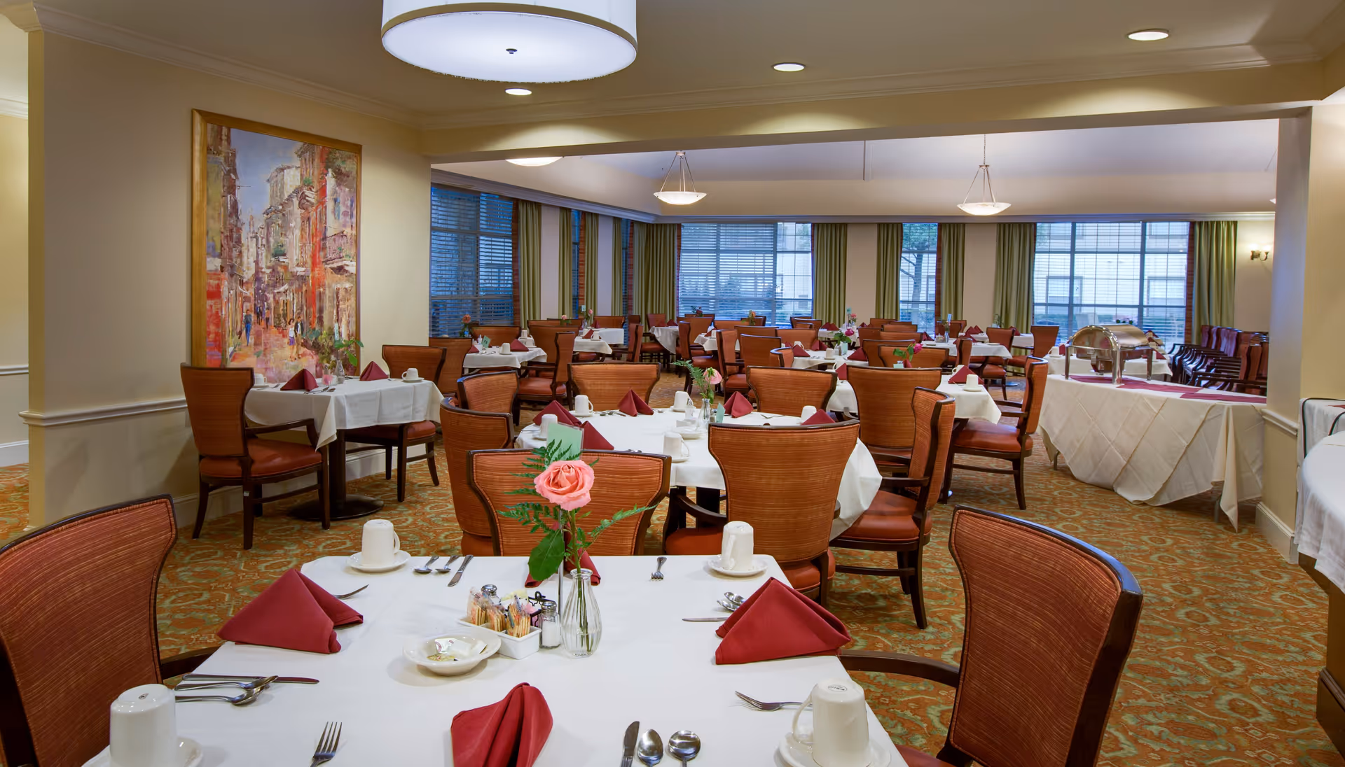 A spacious dining room with multiple tables covered in white tablecloths, each set with red napkins, cups, and silverware. The room features large windows with green curtains, warm lighting from ceiling fixtures, and a colorful painting on one wall. There are also buffet tables covered with white cloths in the background.