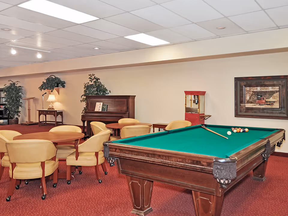 Community lounge with a pool table in the foreground, round tables and chairs, and a piano against the far wall.
