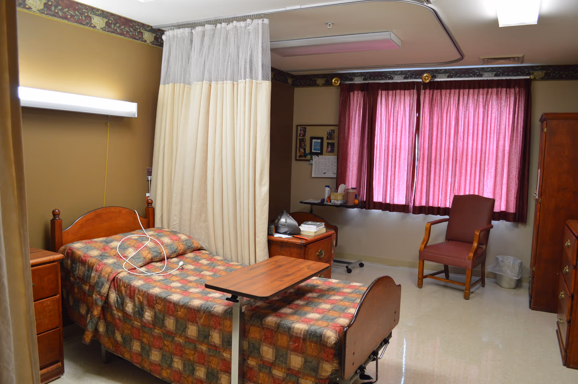 A senior living facility bedroom with a single bed covered in a multicolored checkered blanket. There is a wooden bedside table with a lamp above it, a privacy curtain partially drawn around the bed, a small wooden table on wheels in front of the bed, a wooden chair with armrests near a window with closed red curtains, and a wooden dresser and cabinet along the walls.