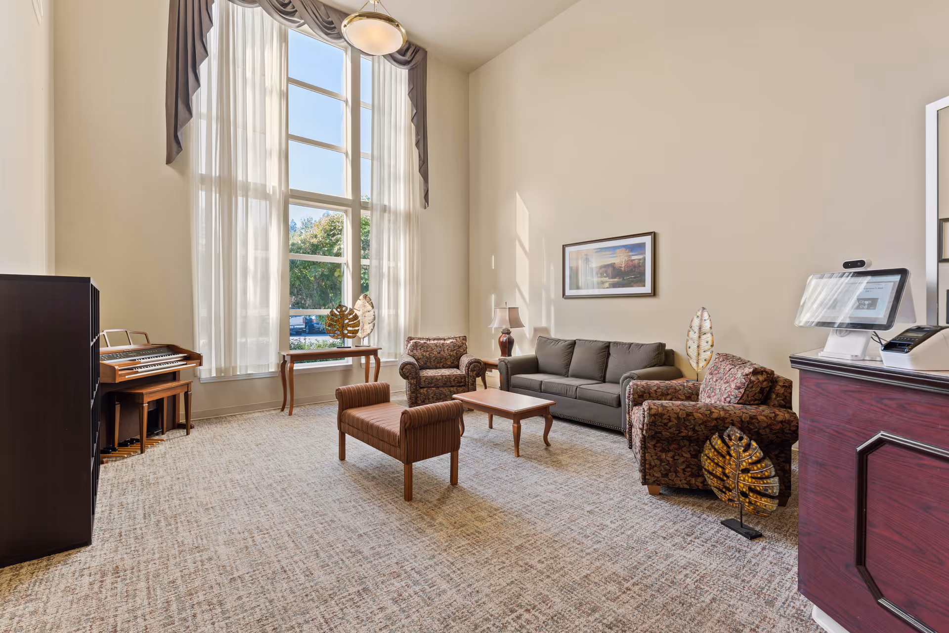 A bright and spacious living room with large floor-to-ceiling windows covered by sheer white curtains and a gray valance. The room features a gray sofa, two patterned armchairs, a wooden coffee table, and a striped bench. There is a wooden piano in the corner and a small table with decorative items near the window. A framed landscape painting hangs on the wall, and a lamp is placed on a side table next to the sofa. The carpet is light-colored with a subtle pattern, and a dark wooden reception desk with a computer monitor is visible on the right side.