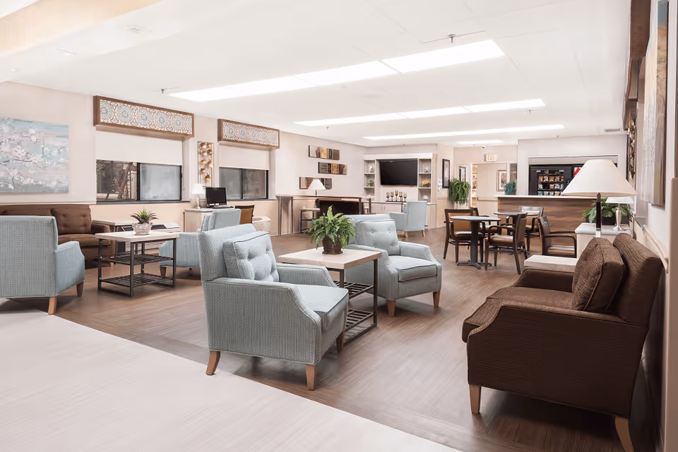 A spacious and well-lit common area in a senior living facility featuring multiple seating arrangements with light blue and brown upholstered chairs and sofas. There are several small tables with plants and lamps, a flat-screen TV mounted on the far wall, and decorative wall art. The room has large windows with patterned valances and a wood floor with a lighter border.