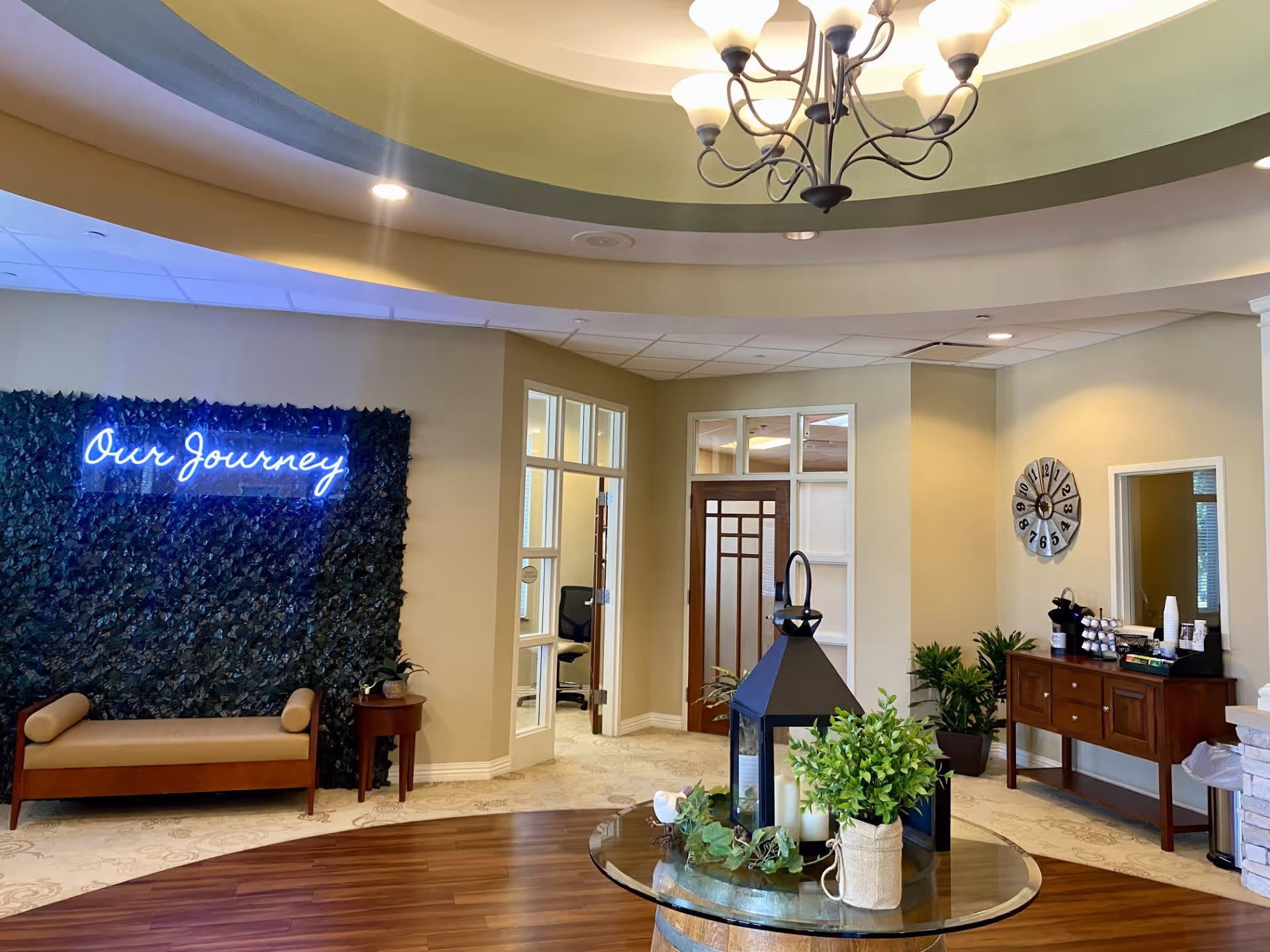 Interior view of a senior living facility lobby area with a circular wooden table in the center decorated with a large lantern and green plants. To the left, there is a bench in front of a wall covered with greenery and a neon sign that reads 'Our Journey'. The room has beige walls, a chandelier hanging from a recessed ceiling, a wooden sideboard with coffee supplies, and a large wall clock.