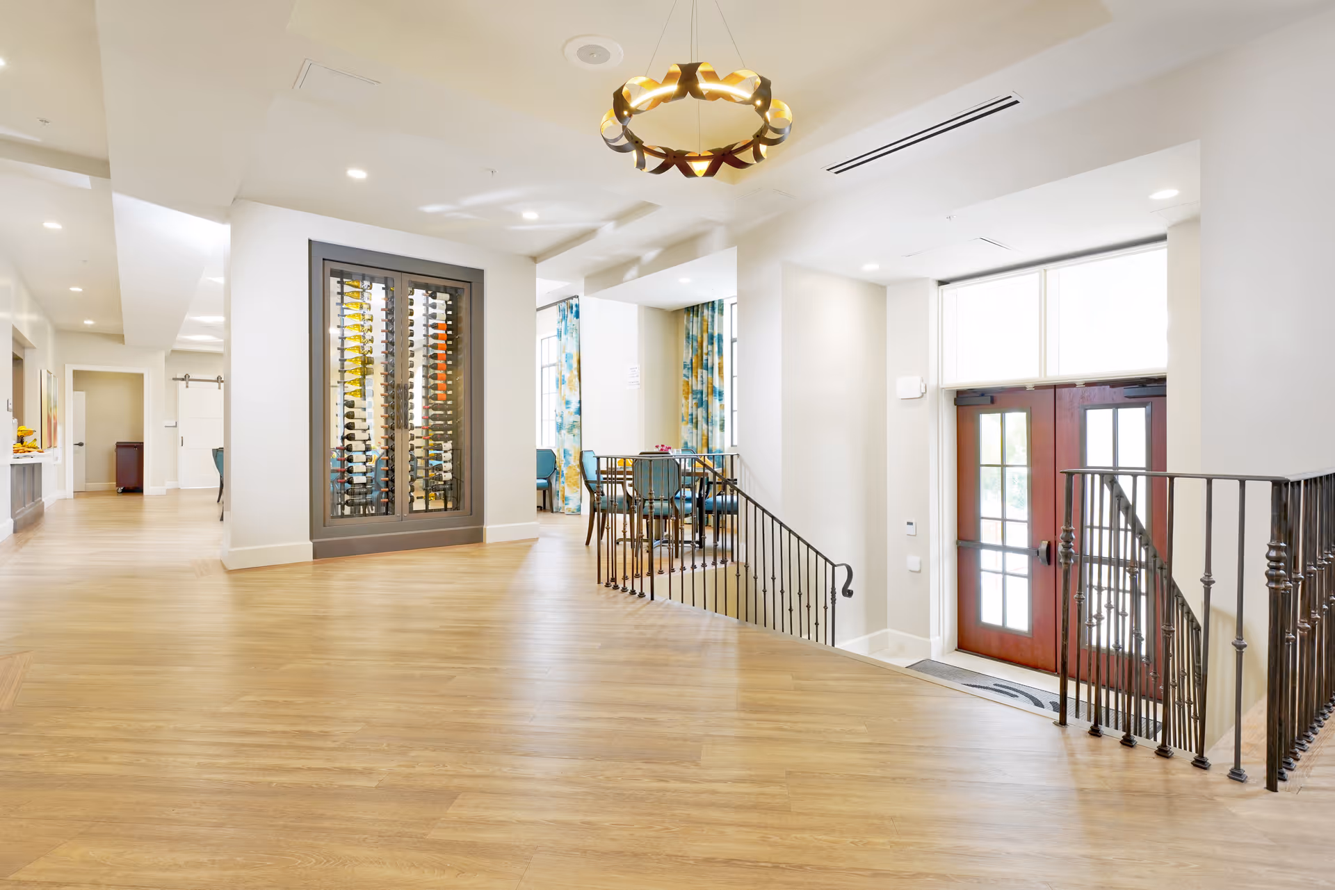 Bright and spacious interior area of a senior living facility with light wood flooring, white walls, a modern chandelier, a wine rack filled with bottles, a small dining table with chairs near large windows with colorful curtains, and a set of double doors leading outside with black metal railings along a small staircase.