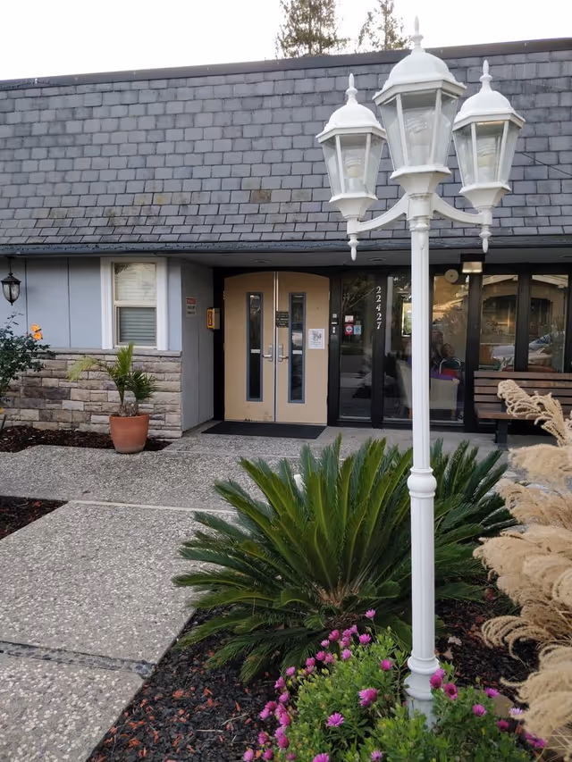Entrance to a building with double beige doors and a glass side panel displaying the number 22427. In front of the entrance is a white three-lantern street lamp surrounded by green plants and purple flowers. The building has a stone and gray exterior with a shingled roof.