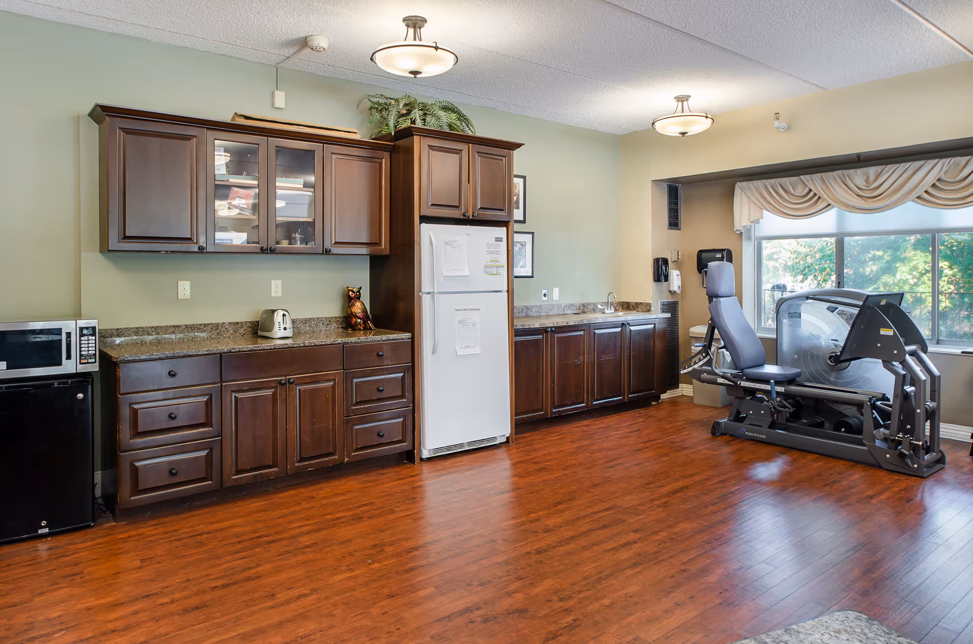Interior room with a dark-wood kitchenette and a seated exercise machine by a large window.
