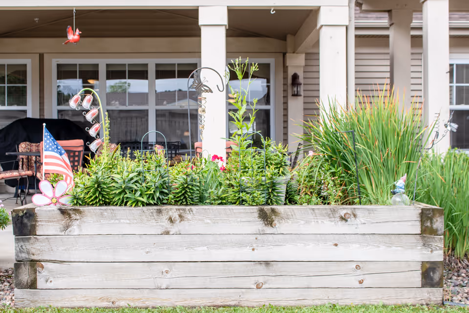 A raised wooden garden bed filled with various green plants and flowers, decorated with a small American flag, a pink flower ornament, and garden stakes. Behind the garden bed is a covered patio area with chairs, a table, and windows of a building.