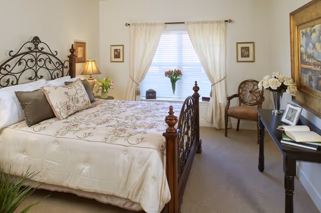 Well-lit bedroom with an ornate wooden bed dressed in floral bedding, a window with curtains, a bedside lamp, and a desk with flowers.