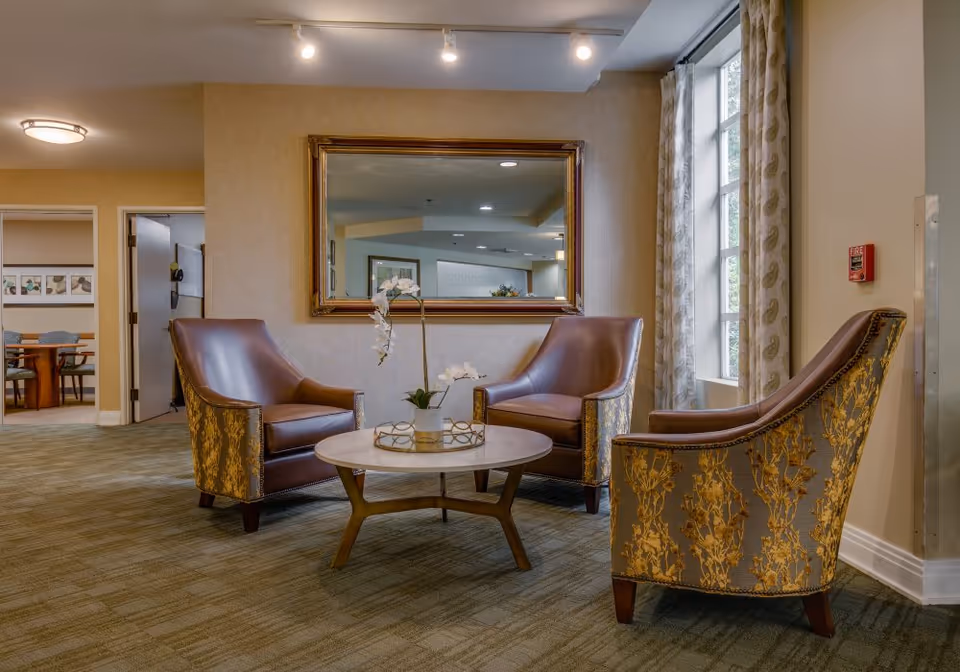 A cozy seating area in a senior living facility with three upholstered armchairs arranged around a round wooden coffee table with a white top. The chairs have a floral pattern on the sides and leather seating surfaces. A large rectangular mirror with a gold frame hangs on the beige wall behind the chairs. To the right, there is a tall window with patterned curtains allowing natural light into the room. The carpet is greenish with a subtle pattern, and in the background, there is a glimpse of another room with a table and chairs.