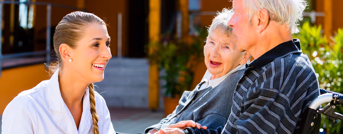 A smiling female healthcare worker with a braid is interacting warmly with an elderly woman and an elderly man in a wheelchair outside a building with orange walls and greenery in the background.