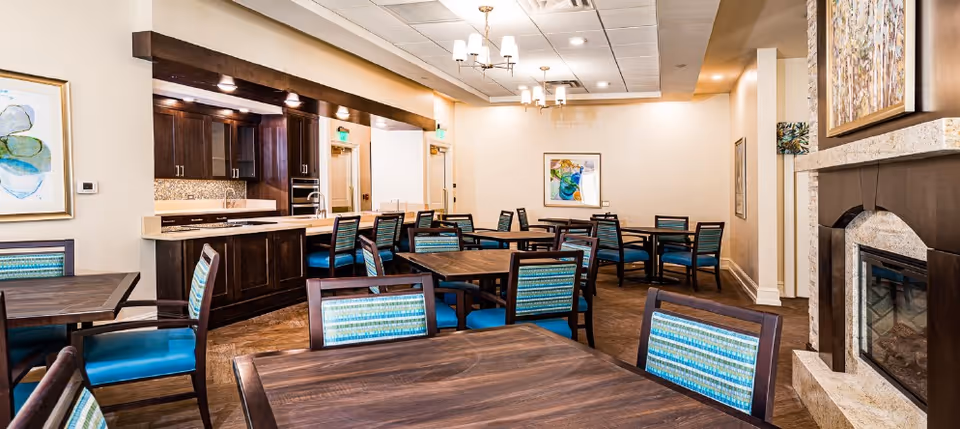 Well-lit dining room with multiple wooden tables and teal-upholstered chairs, a kitchenette area and a fireplace.