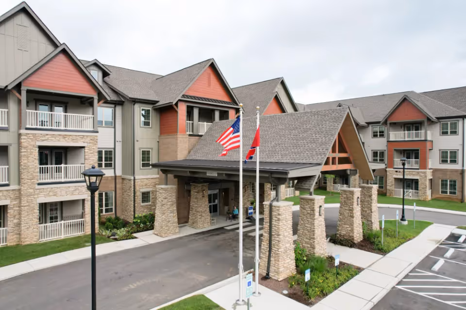 Exterior view of a senior living facility named Harmony At Bellevue featuring a large covered entrance supported by stone pillars, multiple balconies, and an American flag alongside another flag in front of the building.