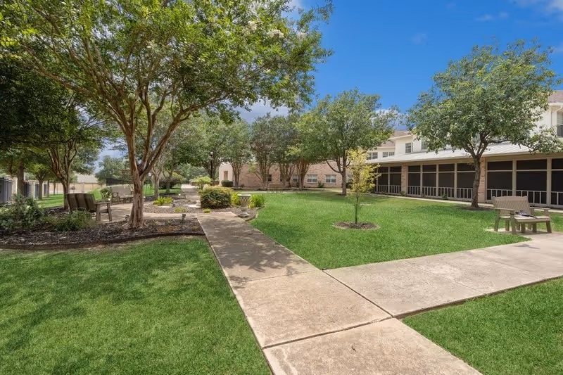 A well-maintained outdoor courtyard area with green grass, several trees, and paved walkways. There are benches placed along the paths, and a building with screened-in porch areas is visible in the background under a clear blue sky.