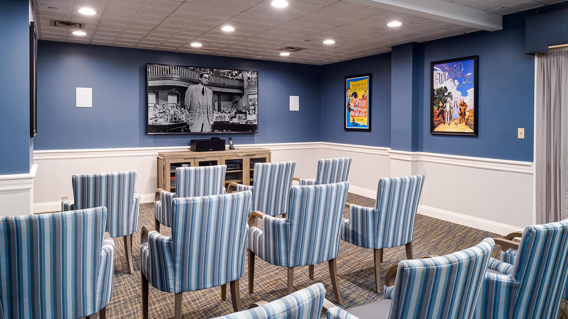 A small theater room with multiple striped upholstered chairs arranged in rows facing a wall-mounted flat screen TV playing a black and white movie. The walls are painted blue with white wainscoting, and there are framed movie posters on the walls.