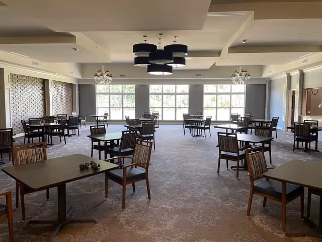 A spacious dining room with multiple square tables and wooden chairs arranged neatly. Large windows along one wall allow natural light to fill the room. The ceiling features modern light fixtures and chandeliers, and the carpeted floor has a subtle pattern.