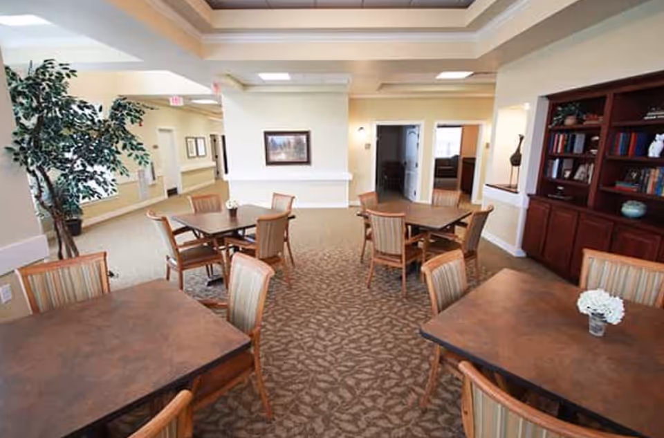 A well-lit common area with multiple square wooden tables and cushioned chairs arranged around them. The room has beige walls, patterned carpet flooring, a large potted plant, framed artwork on the wall, and a built-in wooden bookshelf filled with books and decorative items.