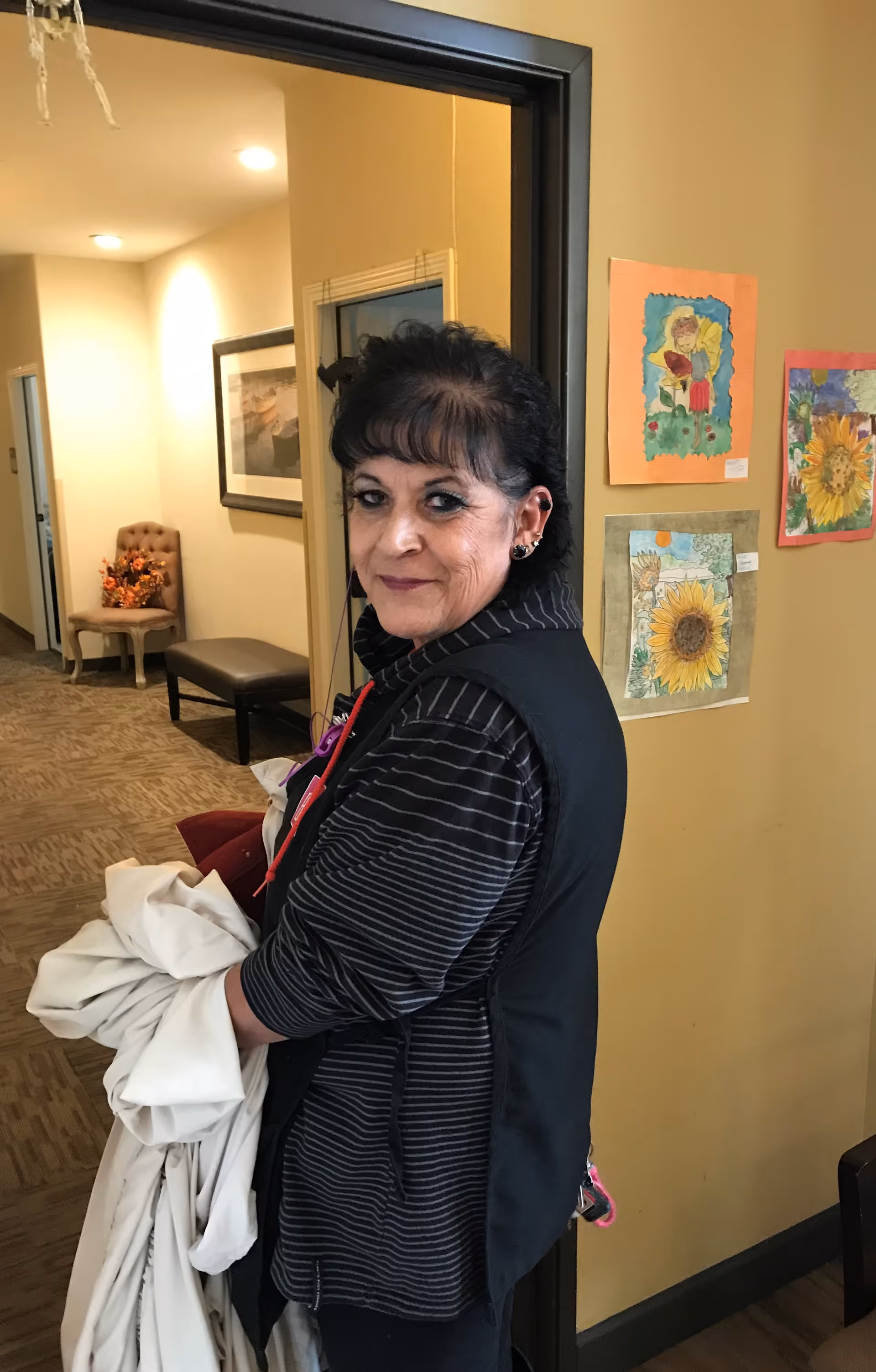 A woman with dark hair and a striped black jacket stands indoors holding a white cloth. Behind her is a hallway with a chair and bench, and a wall decorated with colorful children's artwork featuring sunflowers and other drawings.