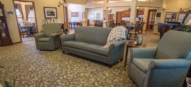 A senior living common area with a sofa and two armchairs on patterned carpet, with dining tables and a service counter visible in the background.