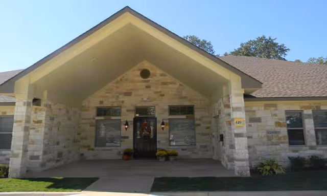 Front entrance of a stone exterior assisted living facility with a covered porch and potted plants.