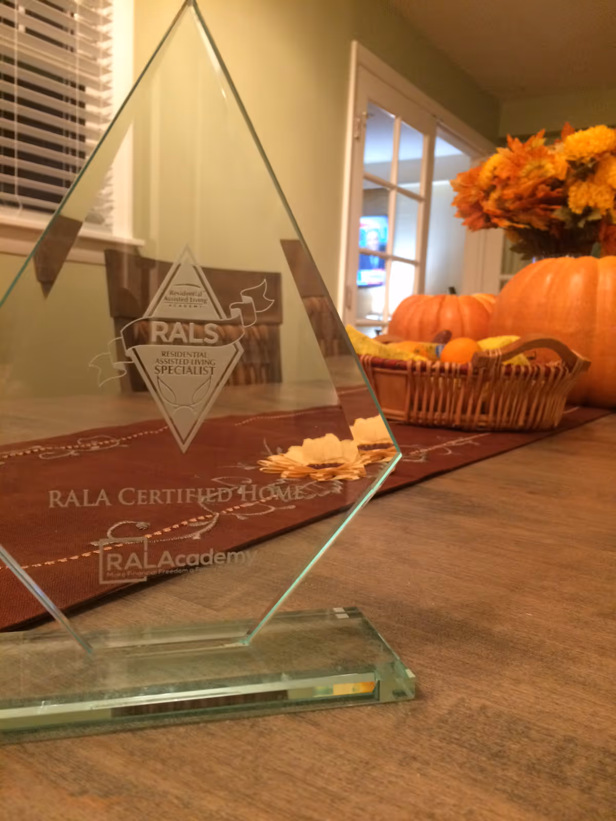 A glass award trophy on a wooden table with a fall-themed centerpiece including pumpkins and flowers in the background. The award reads 'RALS Residential Assisted Living Specialist RALA Certified Home RAL Academy'.