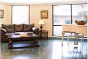 A bright living room with a brown leather sofa, a wooden coffee table, a side table with a lamp, framed artwork on the walls, large windows letting in natural light, and a white grand piano on a tiled floor.
