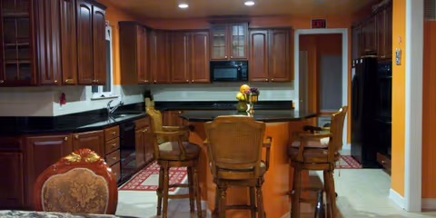 A warm kitchen interior featuring dark wooden cabinets, black countertops, and a central island with three wooden bar stools. The walls are painted orange, and there are rugs on the tiled floor. The kitchen includes a microwave, oven, and sink, with a doorway leading to another room.