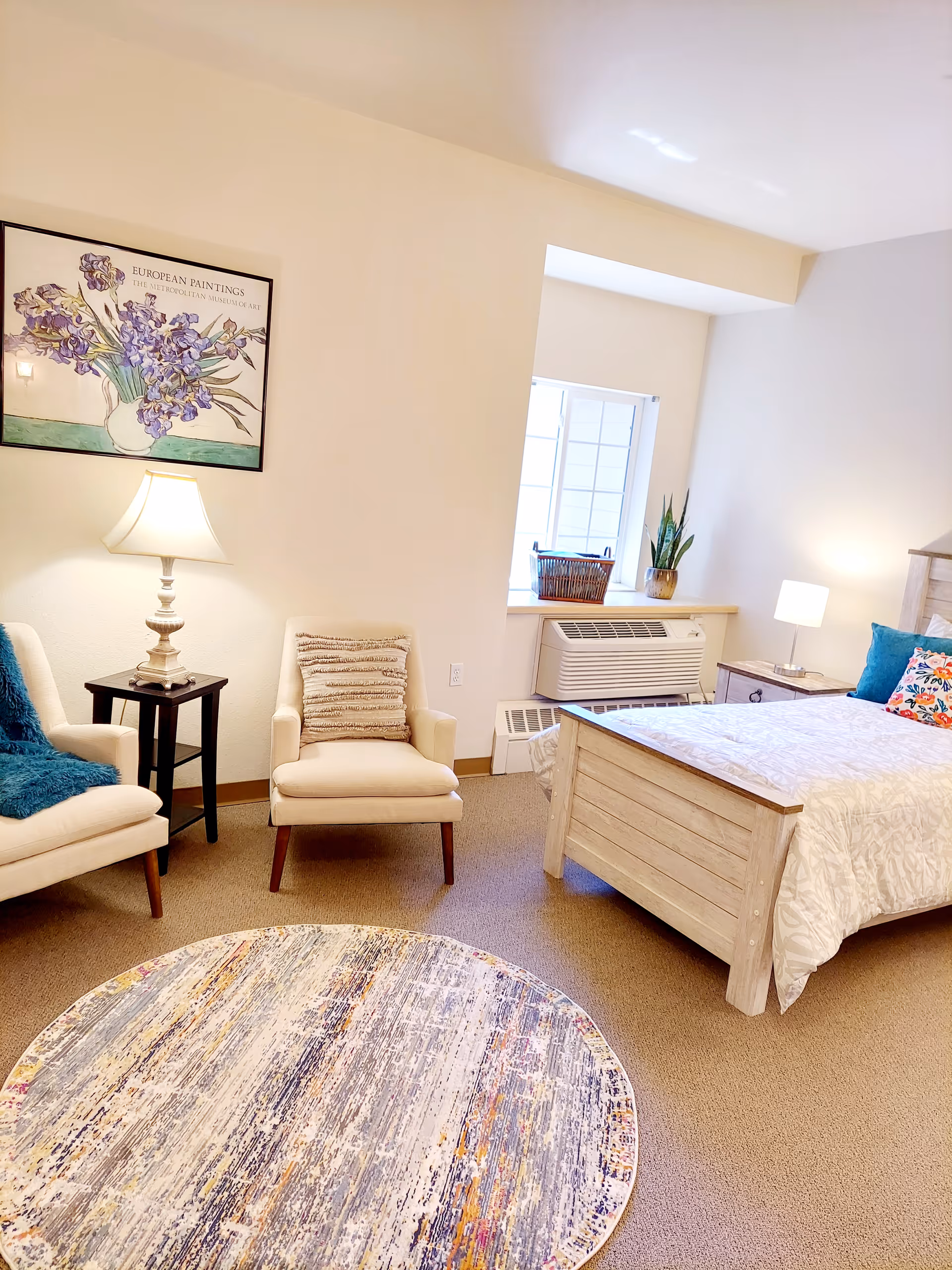 A bright and cozy bedroom with a wooden bed featuring white bedding and colorful pillows. There are two beige armchairs with cushions, a small black side table with a lamp, a round patterned rug on the floor, and a window with a plant and a basket on the windowsill. A framed painting of purple flowers hangs on the wall.
