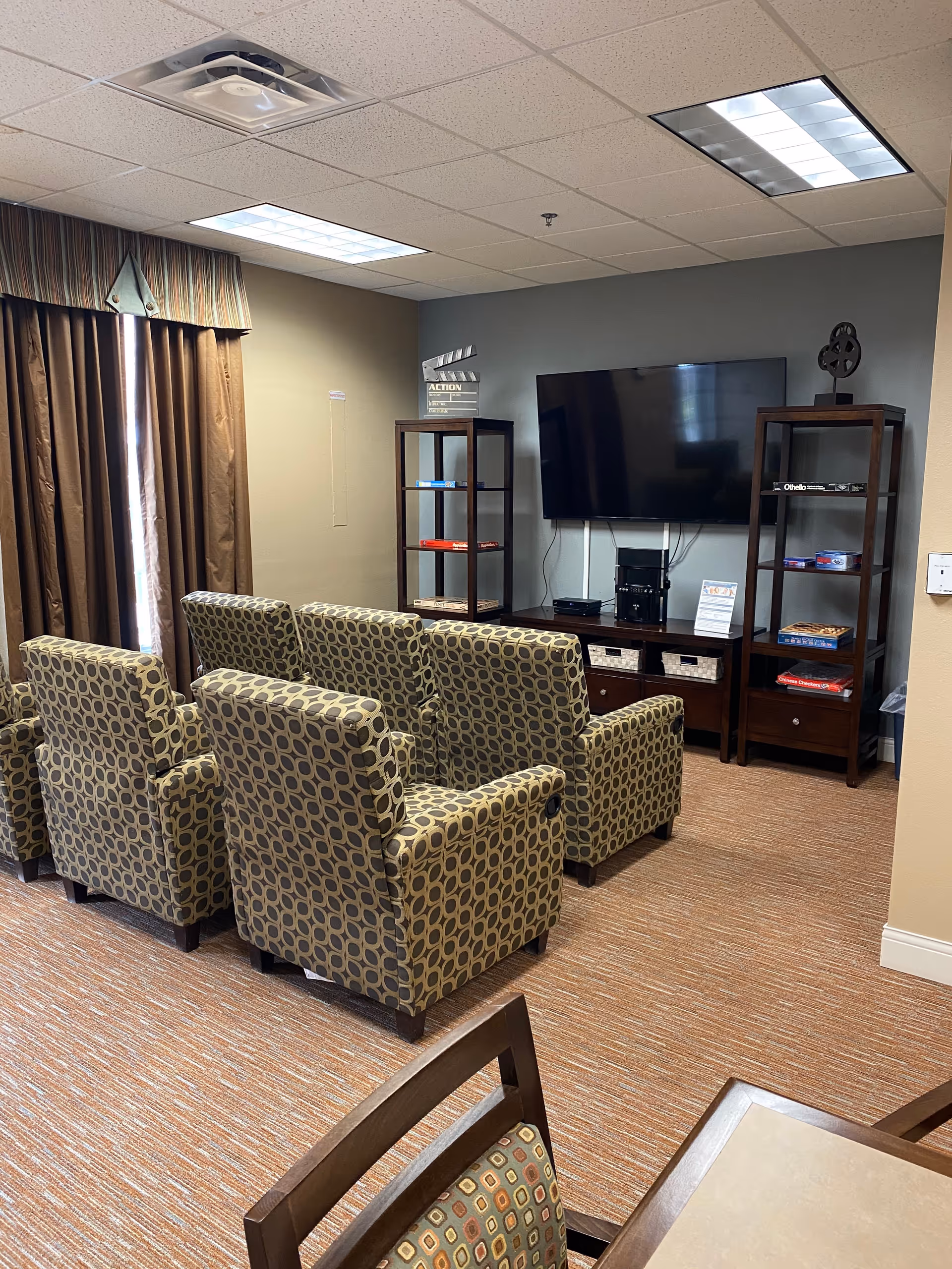 A cozy common area with six patterned armchairs arranged in two rows facing a wall-mounted flat-screen TV. On either side of the TV are tall wooden shelves holding board games and decorative items. The room has brown curtains covering a window and a carpeted floor with a warm tone. A table with chairs is partially visible in the foreground.