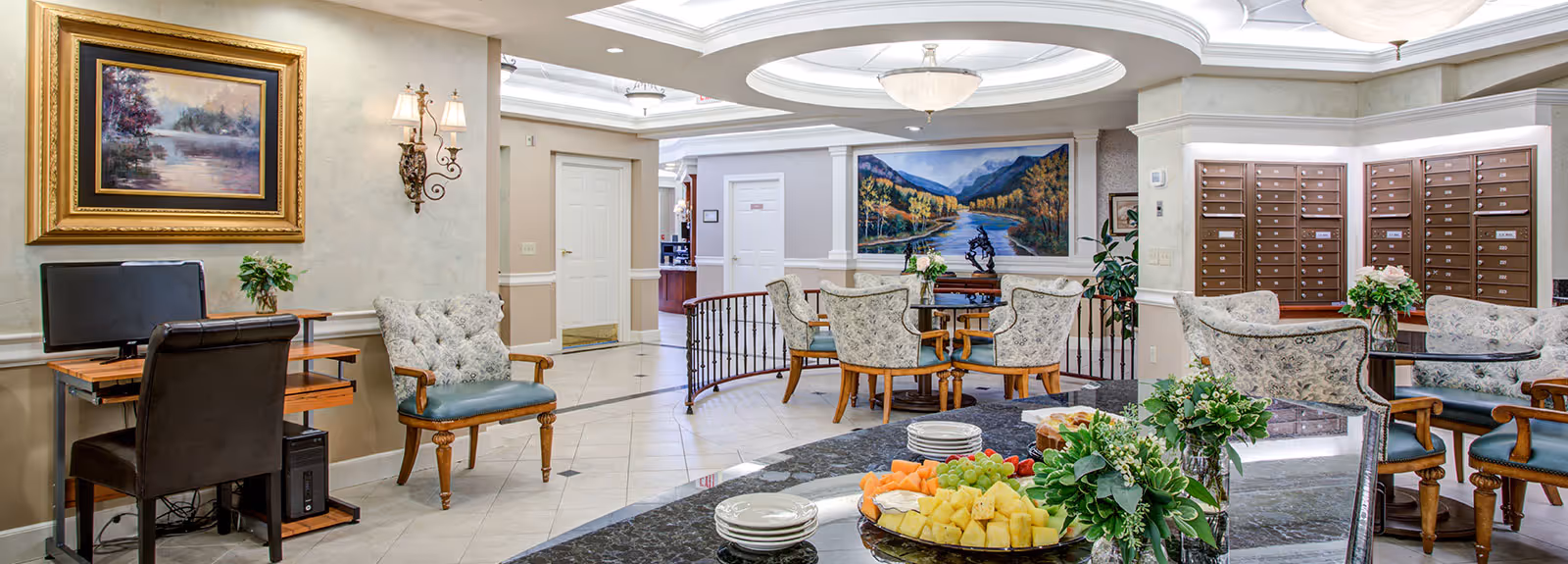Bright communal dining and lounge area with tables and chairs, a marble counter with fruit platters, mailboxes, and wall artwork.