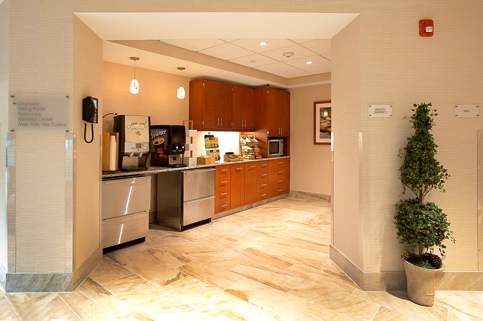 Interior view of a small kitchenette area with wooden cabinets, a microwave, coffee and creamer dispensers, and a countertop with various items. The floor is tiled, and there are two pendant lights hanging from the ceiling. On the left wall, there is a phone and a sign with directions to the courtyard, dining room, restrooms, wellness center, and hair cutters. On the right wall, there are signs for pantry (market) and mail, along with a potted plant.
