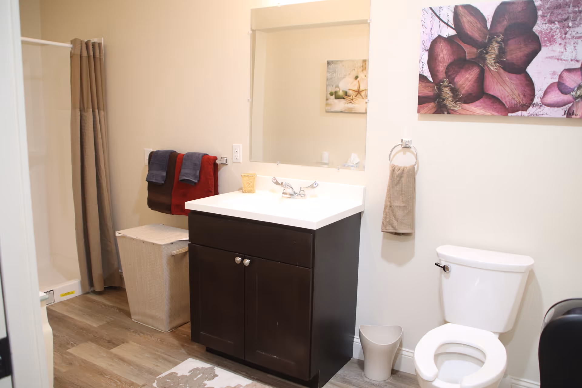 A clean bathroom featuring a white toilet, a dark wood vanity with a white countertop and sink, a large mirror above the sink, a beige towel hanging on a ring, a laundry hamper, and a shower with a beige curtain. There is a floral artwork on the wall above the toilet.