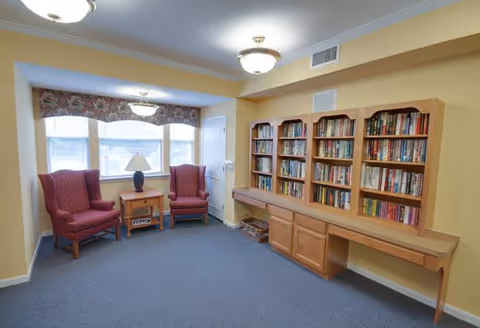 A cozy reading room with two red upholstered armchairs and a wooden side table with a lamp between them, positioned in front of a window with floral valance curtains. On the opposite wall, there are built-in wooden bookshelves filled with books and a long wooden desk below them. The room has beige walls and blue carpeted flooring.