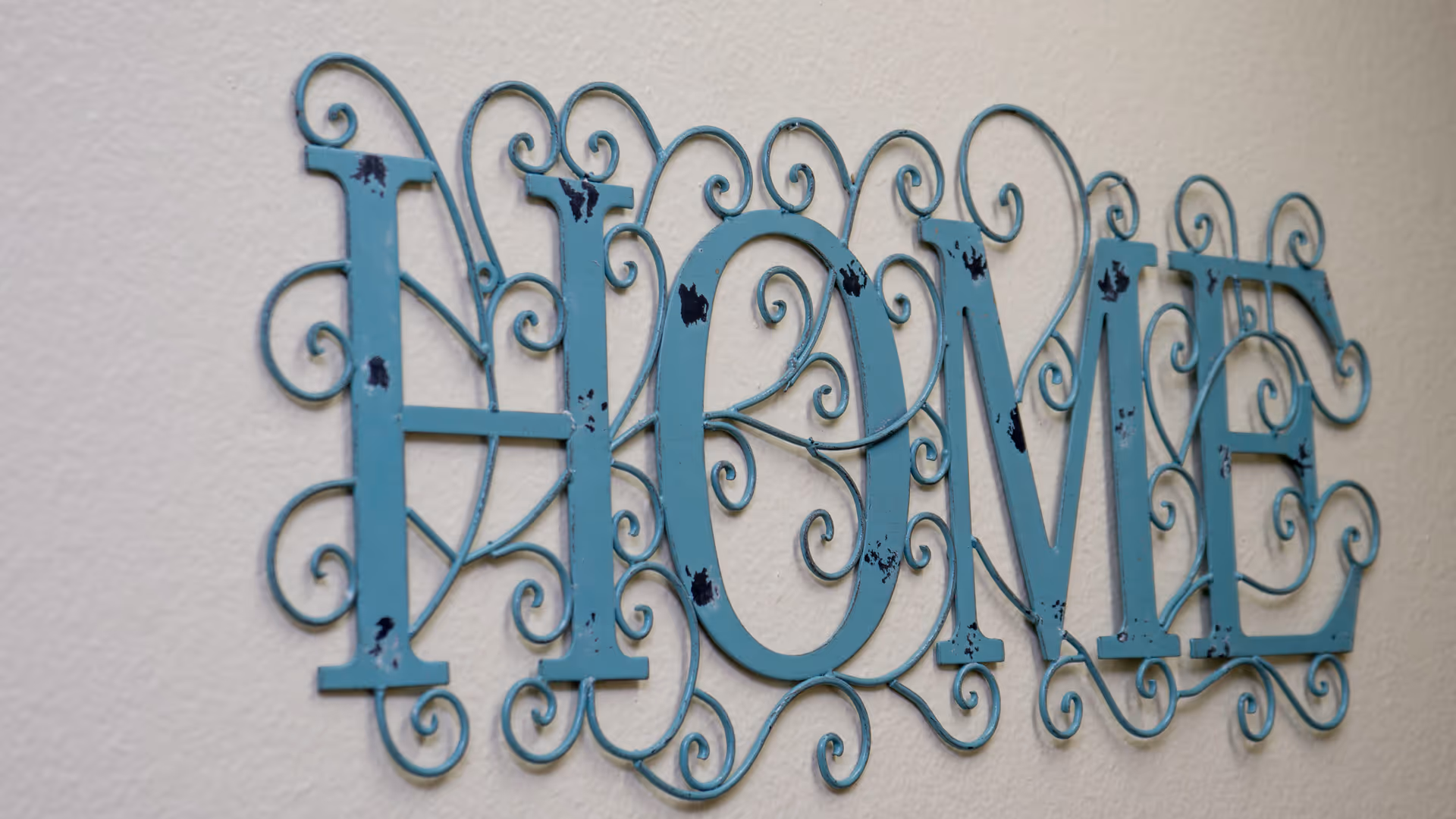 Decorative blue metal wall sign reading "HOME" with ornate scrollwork mounted on a light-colored wall.