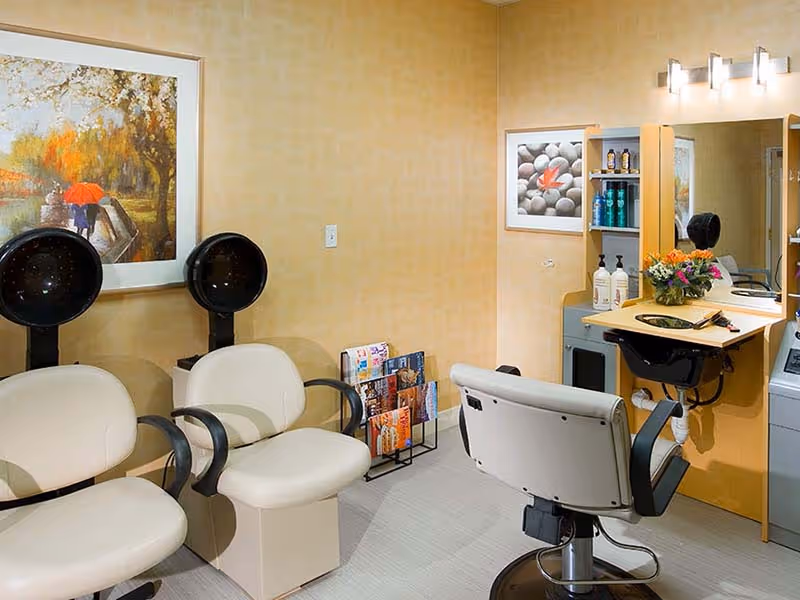 Interior of a hair salon area with two beige salon chairs with black armrests, two black hair dryer stations, a salon styling chair in front of a mirror with shelves holding hair products, a small rack with magazines, and framed artwork on the walls.