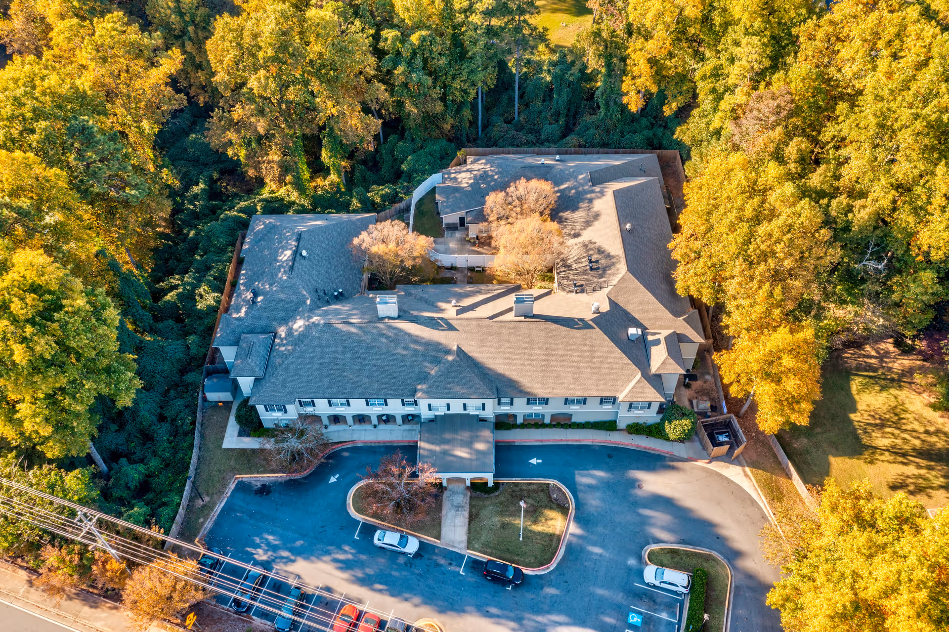 Aerial view of Greenwood Place Assisted Living and Memory Care building surrounded by trees with autumn foliage. The building has a gray roof and a parking lot with several cars parked. The area is lush with greenery and the facility is bordered by a road with visible power lines.