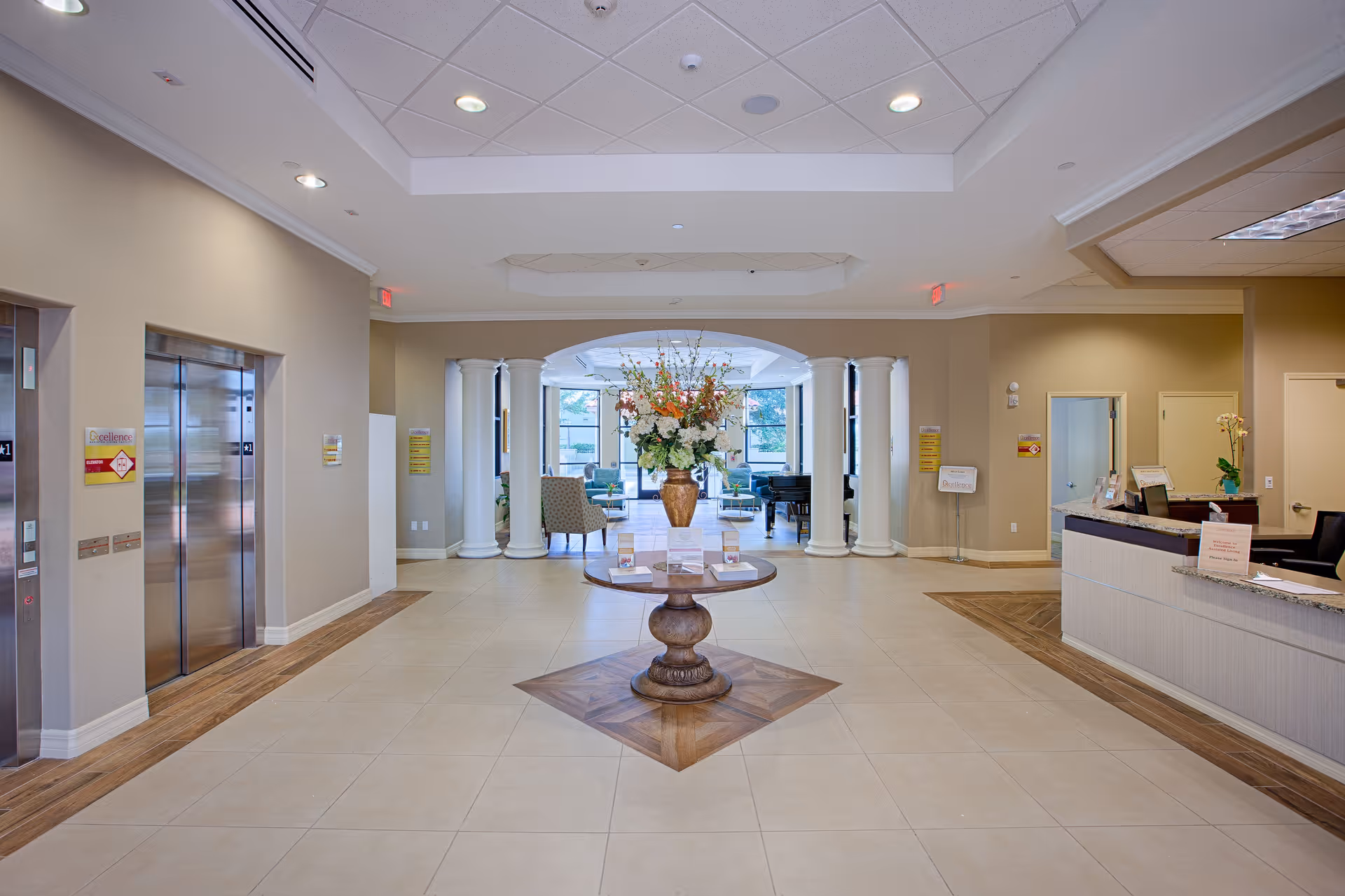 Spacious and well-lit senior living facility lobby with a round wooden table in the center holding a large floral arrangement. To the left are two elevators, and to the right is a reception desk with a computer and some paperwork. In the background, there is a seating area with chairs and a piano near large windows letting in natural light. The floor is tiled with a decorative wooden inlay around the table and along the edges.