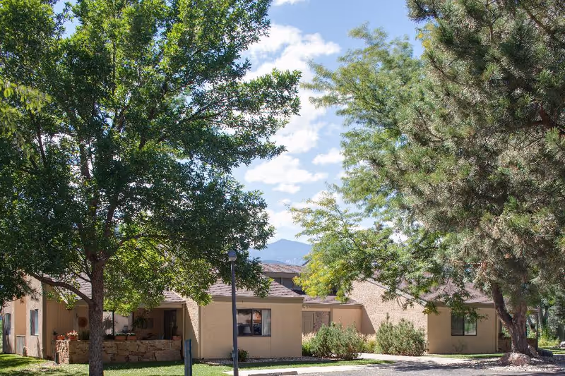 Exterior view of a single-story residential building surrounded by large green trees under a partly cloudy sky with mountains visible in the background.