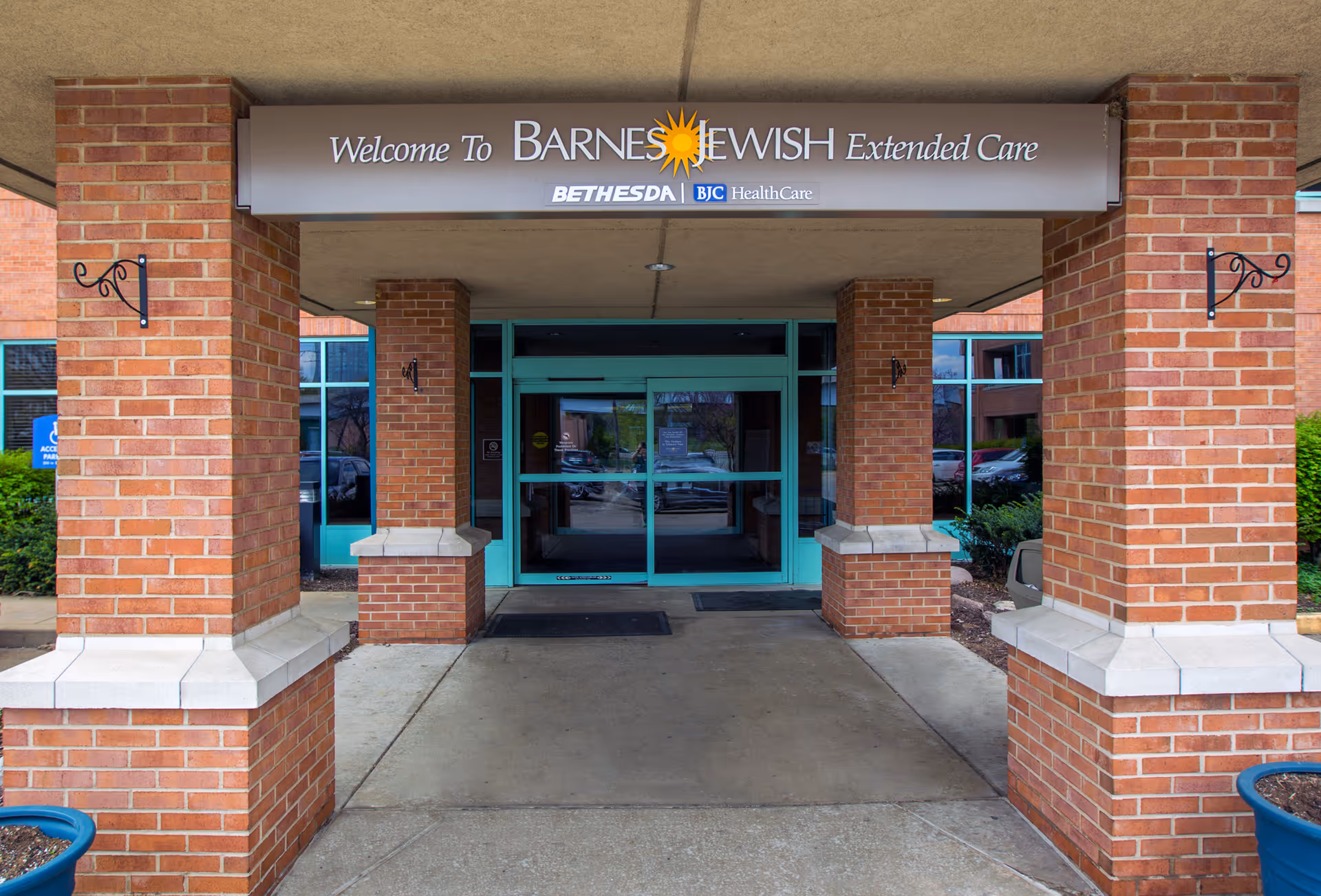 Entrance to Barnes-Jewish Extended Care facility with brick pillars and a covered walkway. The sign above the entrance reads 'Welcome To Barnes-Jewish Extended Care' with Bethesda and BJC HealthCare logos below. Glass doors lead into the building.