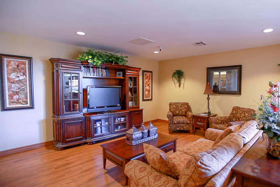 A cozy living room with a wooden entertainment center holding a flat-screen TV, two floral-patterned armchairs, a matching sofa, a wooden coffee table with decorative items, framed floral artwork on the walls, a table lamp, and a potted plant on a side table.