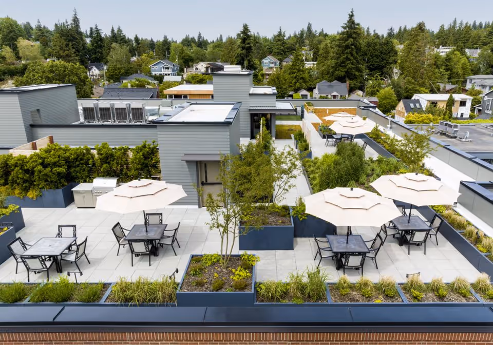 A rooftop patio area with multiple tables and chairs, each shaded by large beige umbrellas. The patio is surrounded by planters with green shrubs and small trees. In the background, there are residential houses and tall trees under a clear sky.